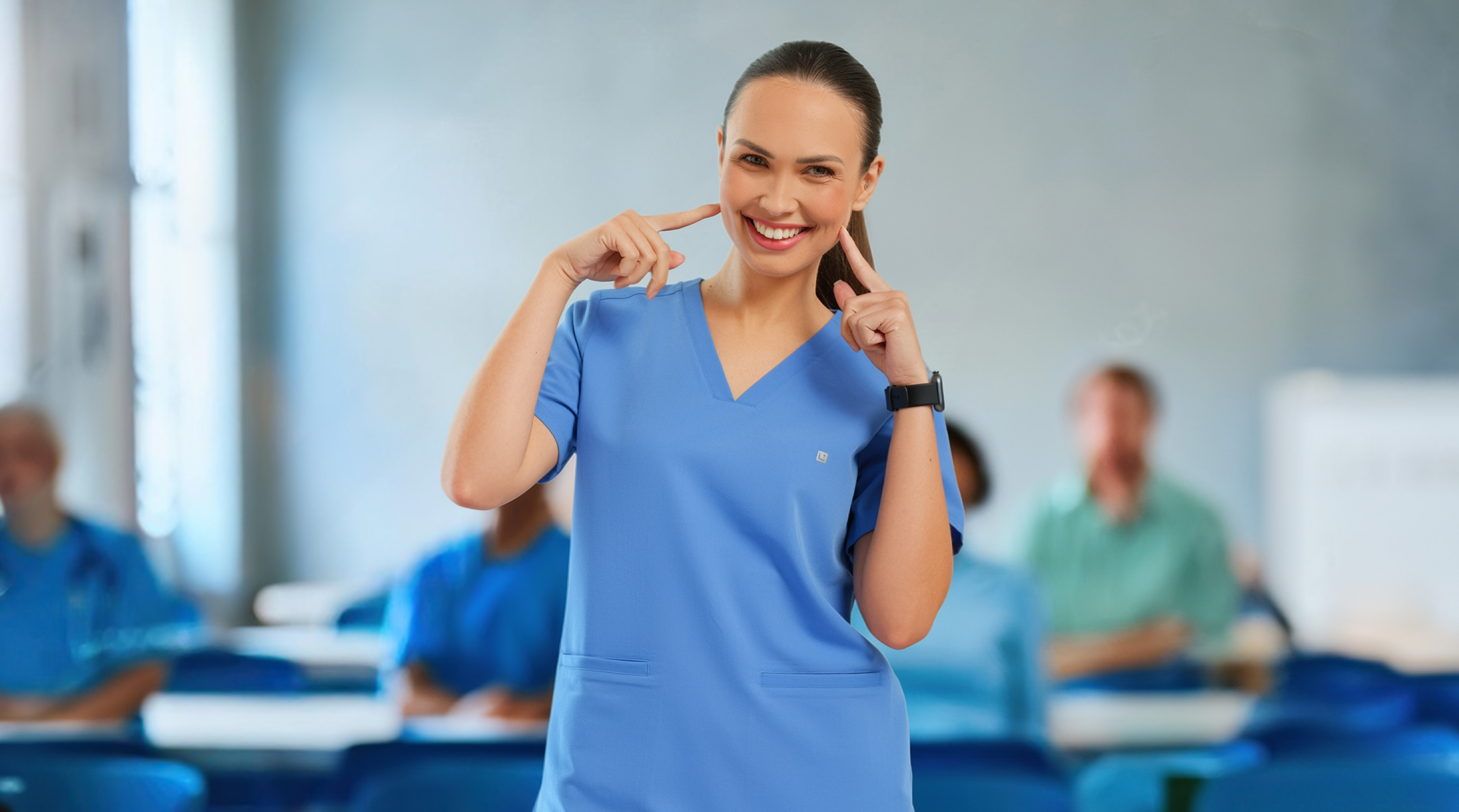 Woman in blue scrubs pointing to her own smile in a classroom setting