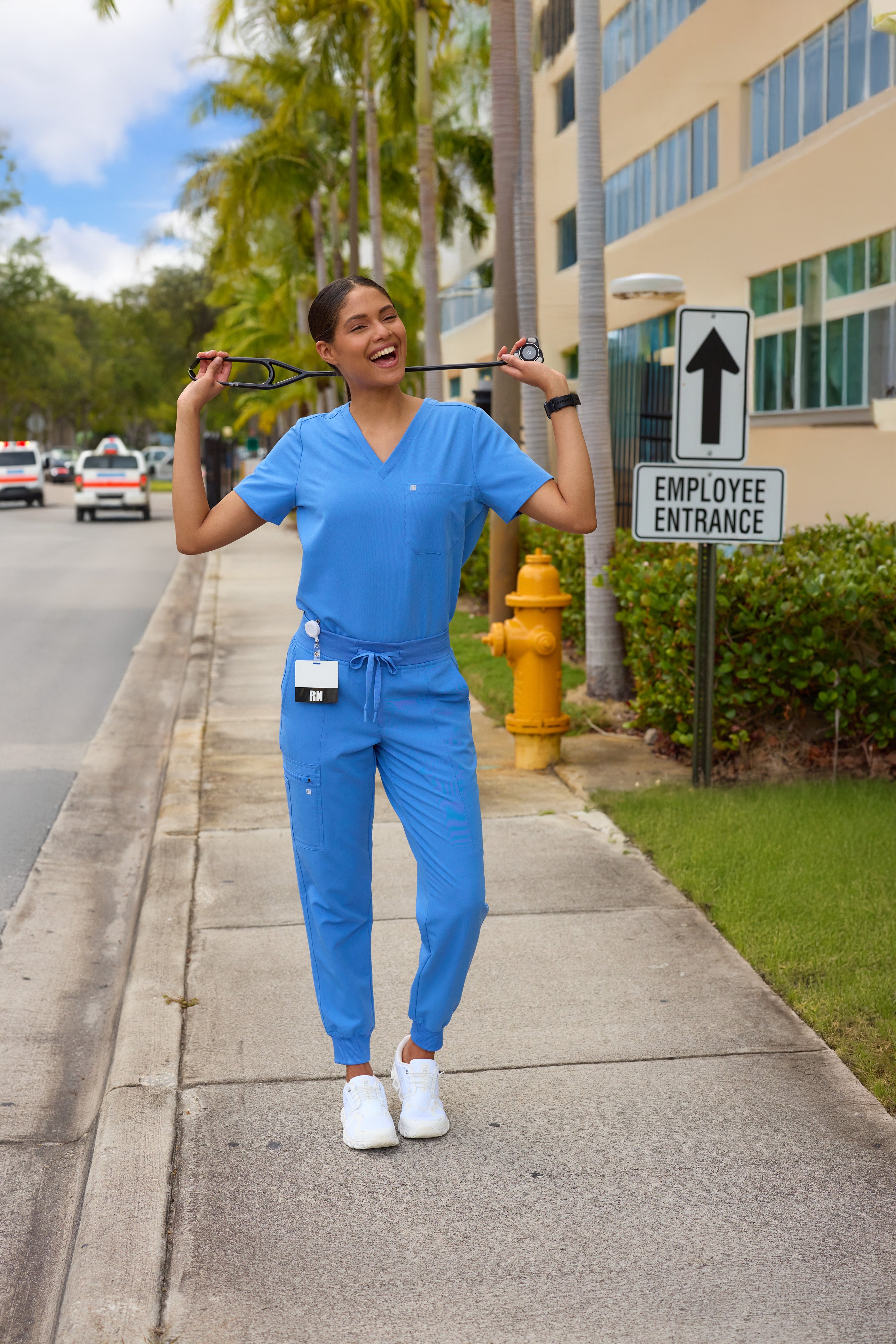 Person in blue scrubs holding sunglasses on a sidewalk with a fire hydrant and building in the background.